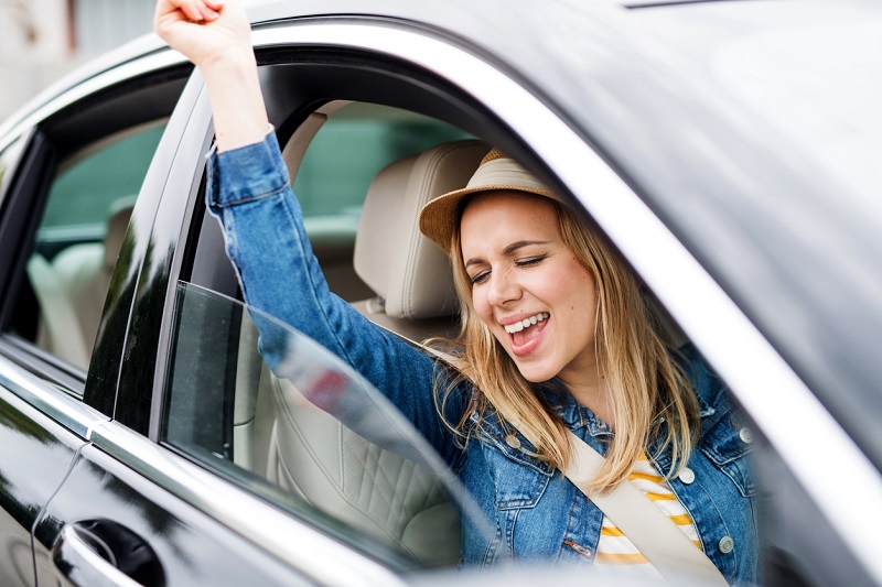A young cheerful woman with hat sitting in car, listening to music and dancing