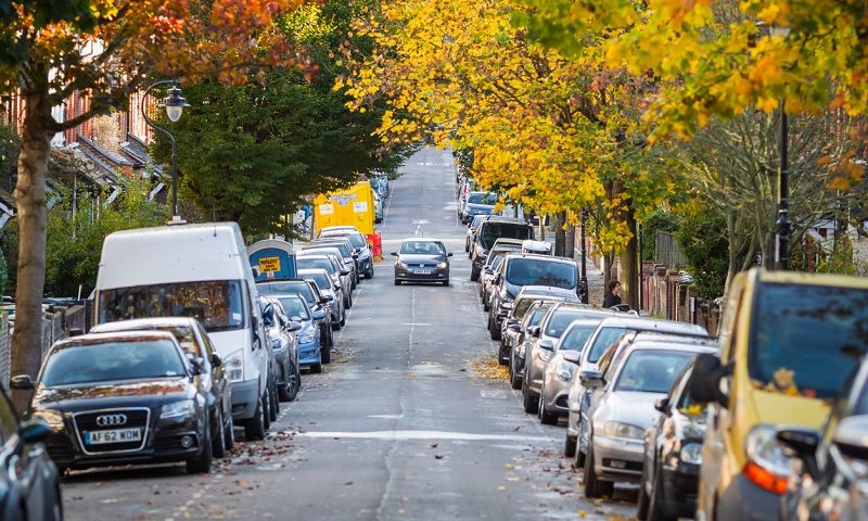A car driving through a treelined street with cars parked on both sides during autumn season in London.